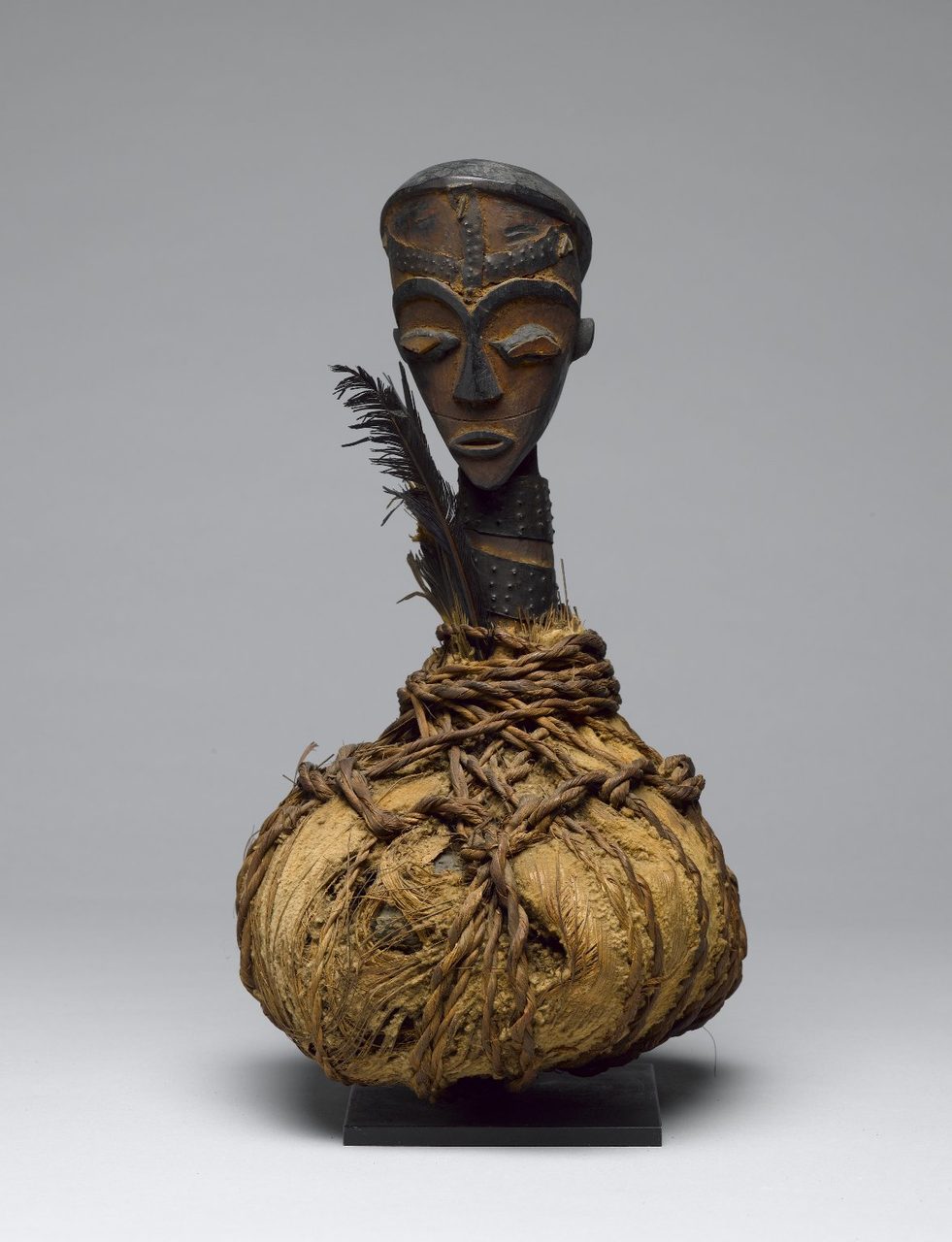 A museum photograph of a boumba bwiti reliquary: a carved wooden head on a long studded neck rising from a large rounded bundle of pale fibre bound with twisted cords, with a single black feather beside the face, on a black mount against a plain grey background