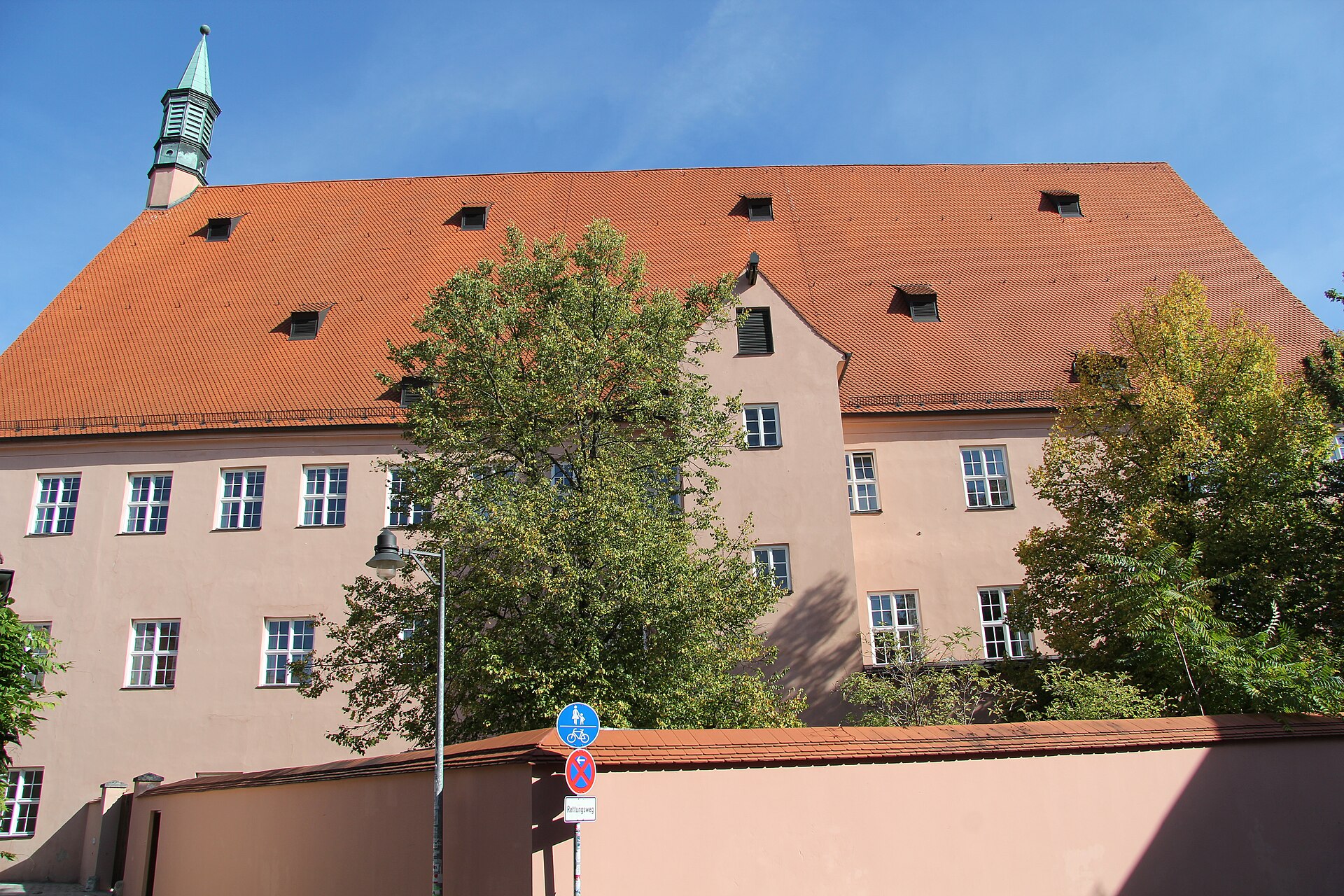 A large salmon-pink rendered building under a steep bright orange tiled roof set with small dormers, photographed from below against a clear blue sky. A slender turret with a green copper spire rises at the left end. Leafy trees stand in front of two rows of white-framed windows, and along the bottom of the frame run a low rendered wall, a modern streetlamp, and a post carrying German road signs including a no-stopping sign.