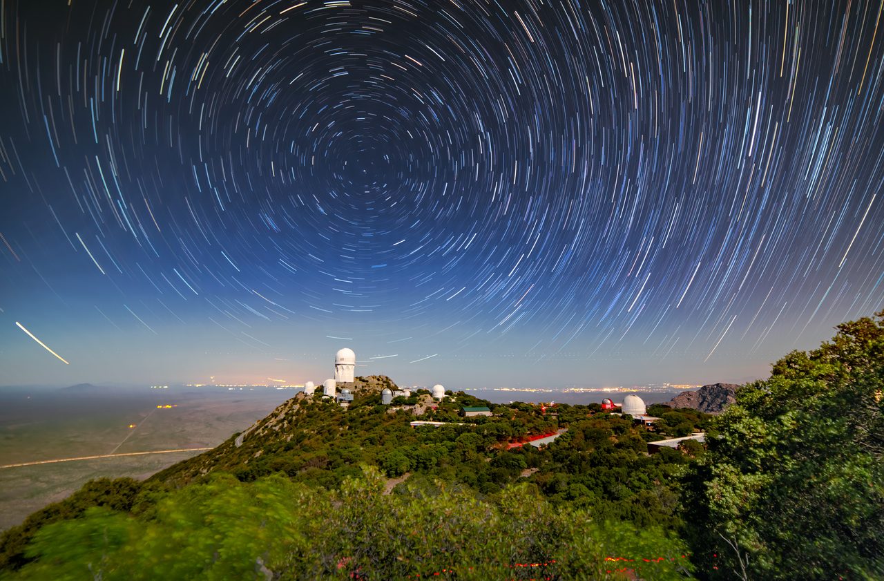 A long exposure photograph in which the stars above Kitt Peak National Observatory are drawn into complete concentric circles around a still point, with several observatory domes on the wooded ridge below and a band of city light along the horizon