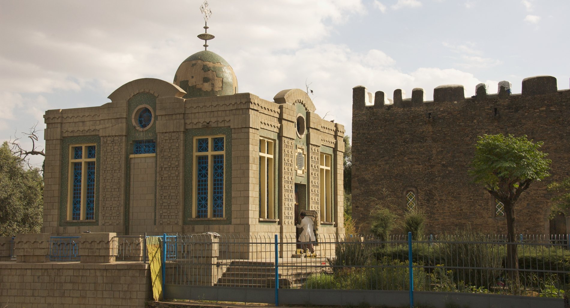 A modest rectangular chapel in pale block masonry with green decorative panels and tall cruciform windows holding blue geometric leaded glass. A patched green dome carries an ornate Ethiopian cross finial. A single figure in a white robe stands at the dark doorway. A metal railing fence with blue posts and a yellow gate rings the whole building. An older crenellated stone wall stands to the right.