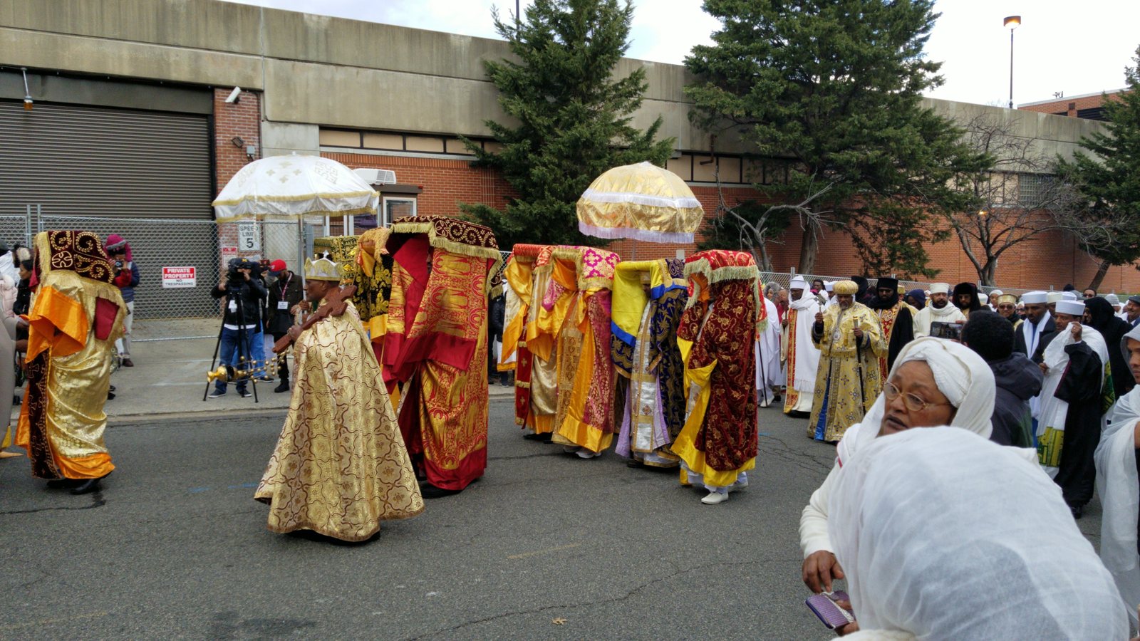 A religious procession. Priests in elaborate gold, crimson, pink, blue and yellow brocade vestments, several wearing crowns, carry objects on their heads that are completely wrapped in embroidered cloth. Two fringed ceremonial umbrellas, one white and gold, one gold. A crowd in white shawls at the right. Behind them a chain-link fence, a brick warehouse, a concrete parking structure and signs reading Private Property No Trespassing and Speed Limit 5.