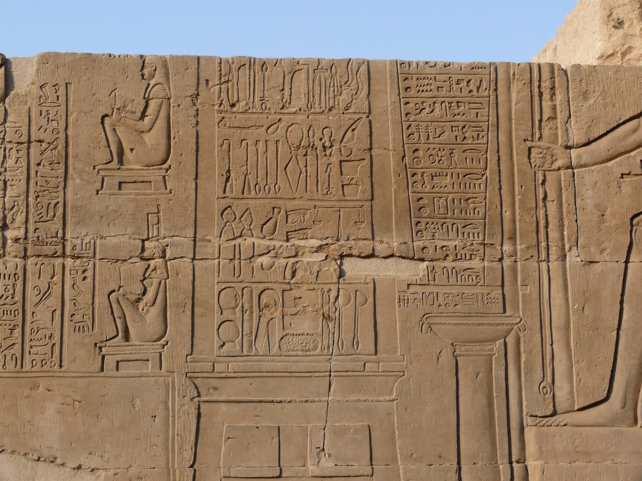 A carved temple relief showing two registers of surgical instruments filling the center, two smaller seated figures at the left, and columns of hieroglyphic text at right
