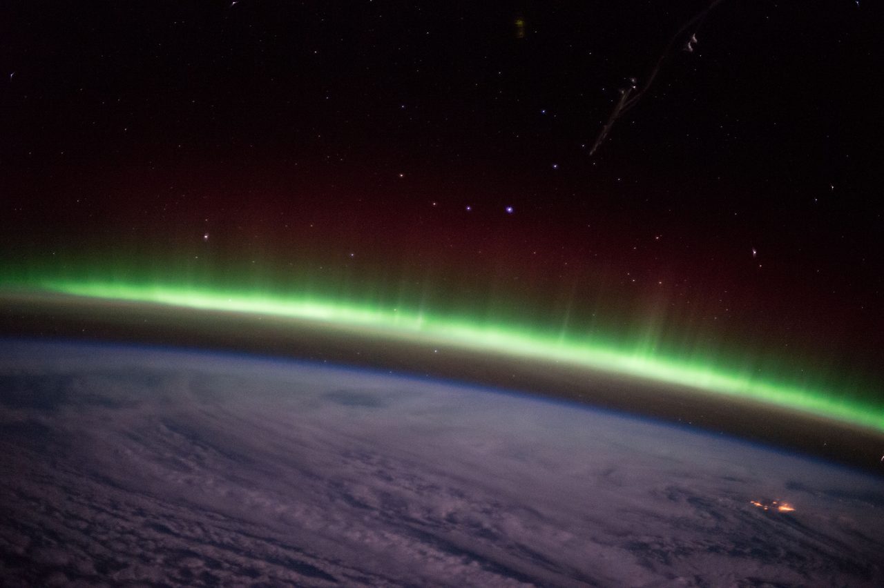A green auroral band curving along Earth's limb photographed from orbit, with a dark red upper-atmosphere glow above it, stars overhead and cloud-covered ocean below