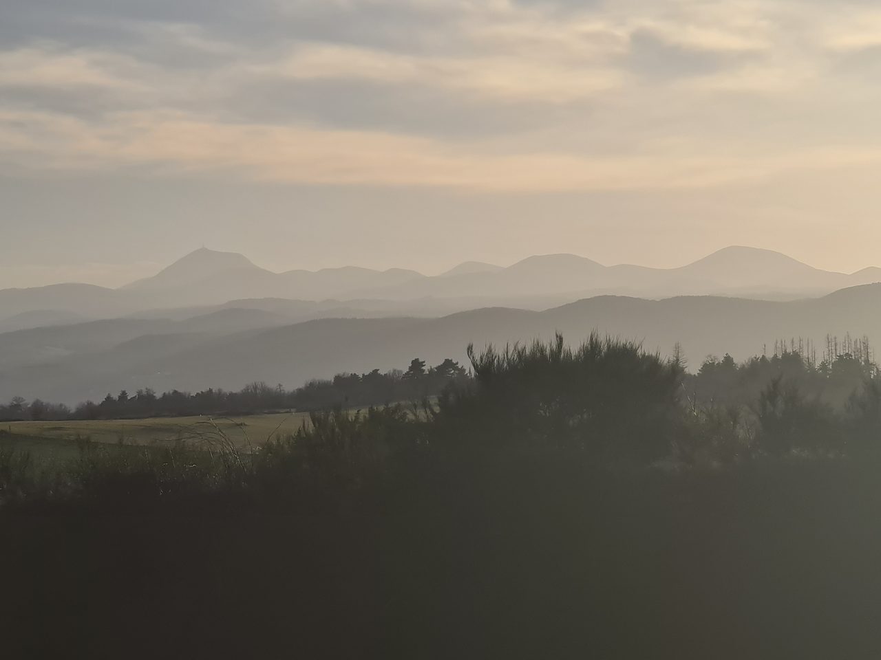 A hazy backlit panorama of the Chaine des Puys, the volcanic cones reduced to layered grey and blue silhouettes receding into mist under a pale sky, the tallest summit at left carrying a slender mast, with dark scrub and trees across the foreground