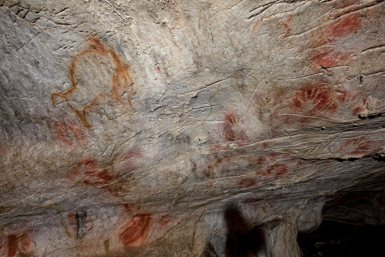 A cave wall at El Castillo covered with red hand stencils of varying size, with an outlined ochre bison at upper left, across pale fissured limestone