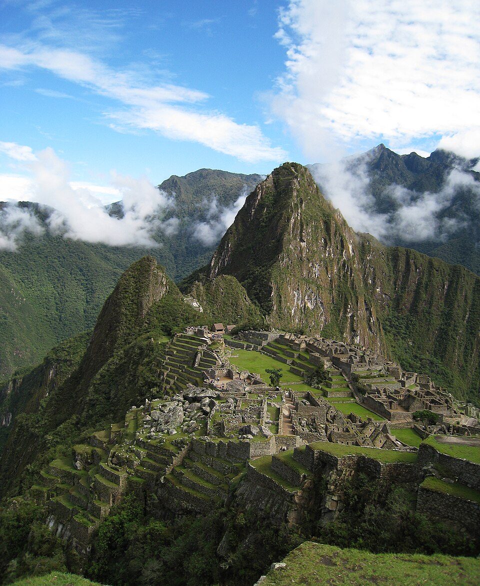 An elevated overlook of the full Machu Picchu urban sector on its ridge, terraces stepping down both flanks, with Huayna Picchu behind it and the valley falling away on both sides
