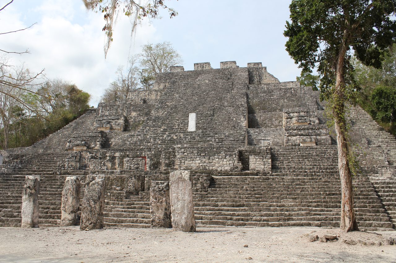 The broad stepped mass of Structure II at Calakmul rising behind a long flight of stairs, with a row of five weathered standing stones set along the base of the steps and forest crowding both sides