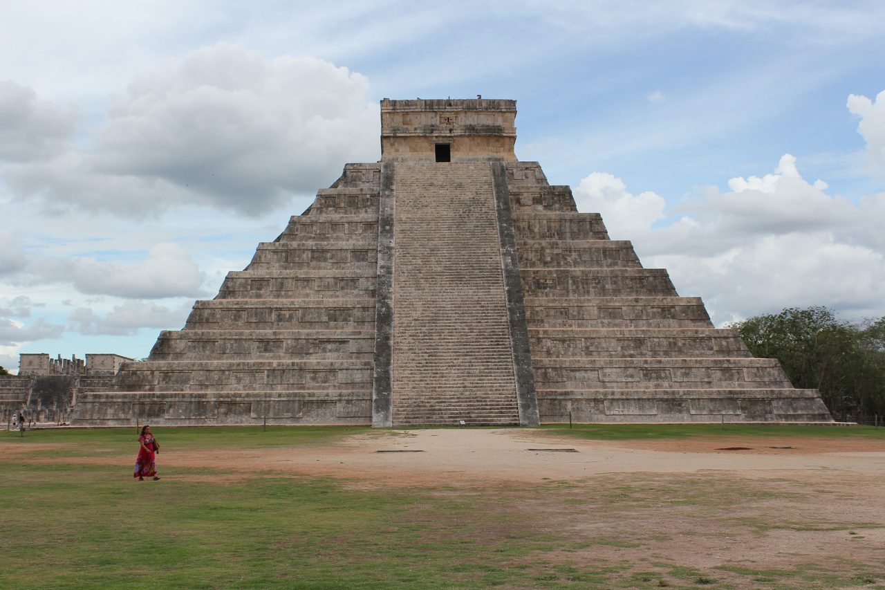 El Castillo at Chichen Itza seen from the west, a nine-terraced stone pyramid with a central stairway and a temple on top, under a bright clouded sky, with a woman in a red patterned dress walking across the grass at lower left for scale