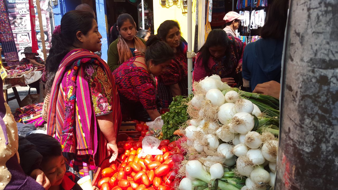 A crowded market aisle photographed close up: women in brightly woven huipiles buying and selling, heaps of tomatoes and white onions with their roots still on in the foreground, textiles hanging at the stalls behind, and a child at the lower left