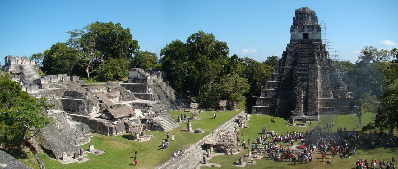 A wide view across the Great Plaza at Tikal: a steep stepped temple at right with scaffolding up one side, the terraced North Acropolis at left under modern thatch shelters, mown grass between them, and a crowd of people gathered at lower right around a smoking fire