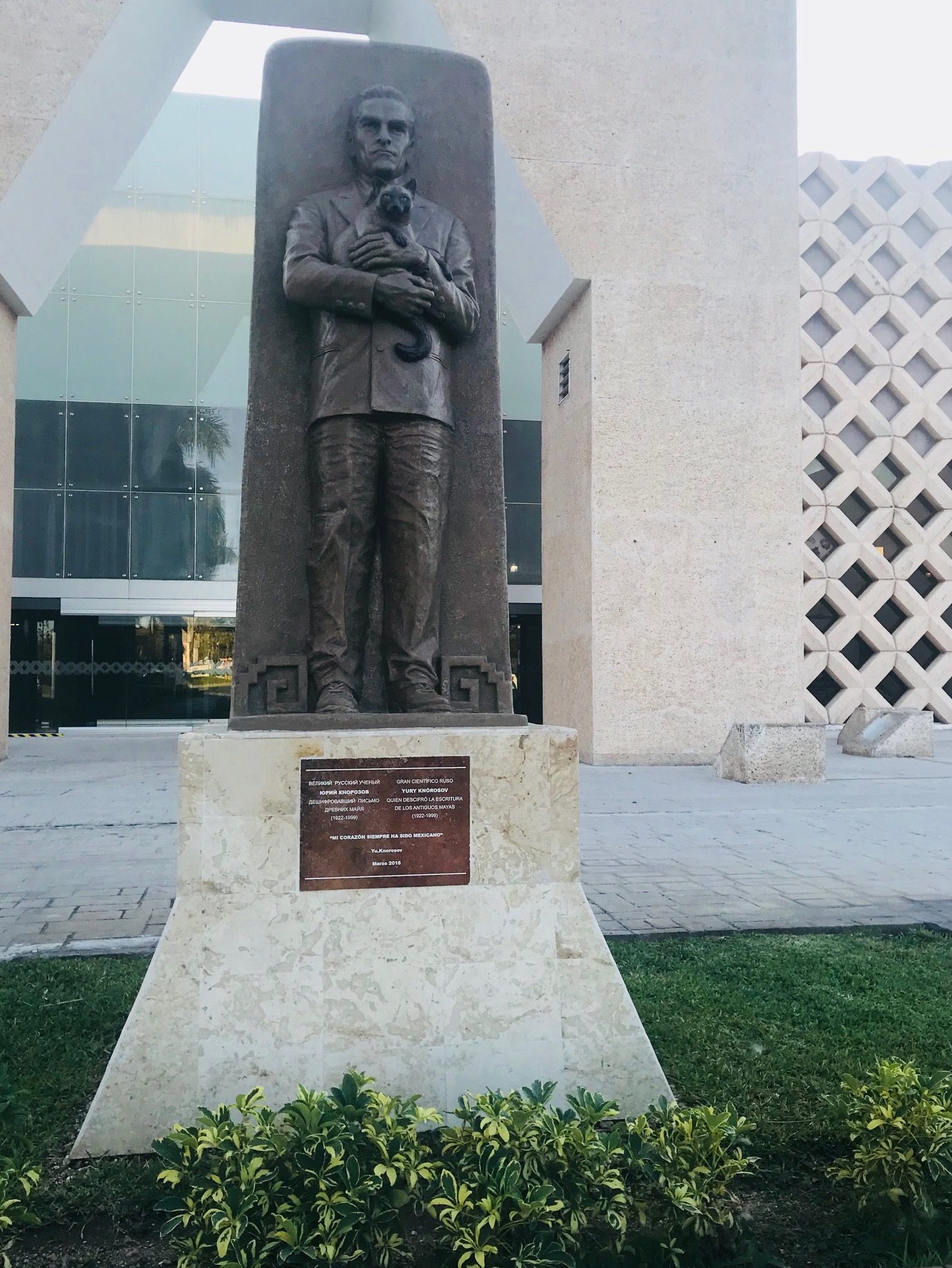 A bronze statue on a pale stone plinth outside a low modern museum building, shot in flat evening light. A man in a jacket and trousers stands against a tall bronze slab, holding a small cat against his chest with both hands, its tail curling down. A stepped fret pattern is cut into the slab near his feet. A dark plaque on the plinth carries text in Russian and Spanish. Clipped shrubs run across the foreground.