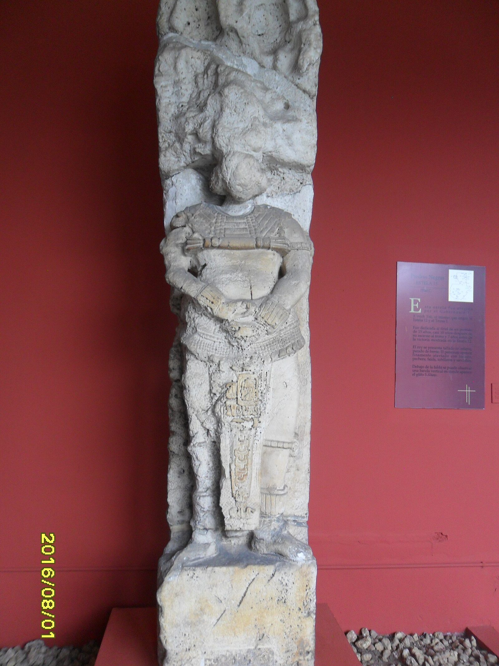 A tall, narrow limestone monument standing against a deep red museum wall. Carved in high relief is a standing human figure in elaborate costume, wearing a broad pectoral, a patterned skirt and heavy anklets, with arms held across the body. The stone is heavily eroded above the waist and the head is largely lost. A vertical band of small carved blocks runs down beside the figure's skirt. A purple museum label panel hangs on the wall to the right. A yellow camera date stamp reads 2016/08/01 in the lower left corner.