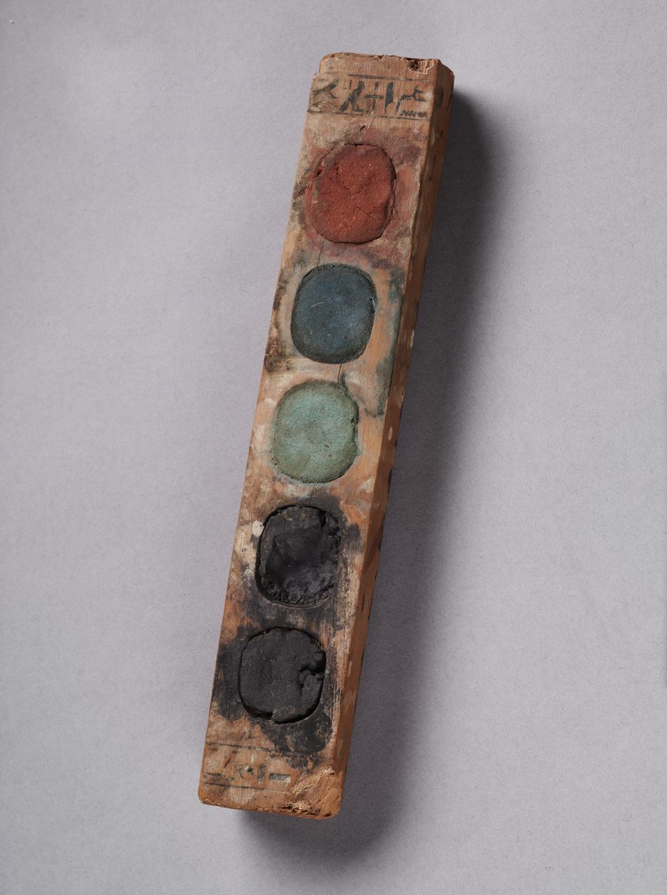 A long, narrow wooden bar photographed against a plain museum ground, lying at a diagonal from the upper right to the lower left with a soft shadow beneath it. Five oval wells are cut in a single row down its upper face, each holding a rounded, cracked cake of dried pigment, and a short line of hieroglyphic ink inscription runs across the wood at each end. The bar is worn, stained and splintered at both tips.