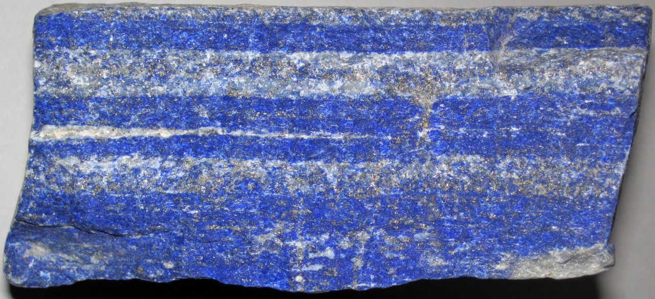 A single flat-cut slab of banded rock lying on a plain, evenly lit ground and filling almost the full width of a wide, shallow frame, with a dark shadow along its lower edge. The cut face is crossed by roughly horizontal bands of coarser, lighter mineral running through a dense granular mass, fine metallic flecks are scattered across the whole surface, and both ends of the slab are broken and irregular.