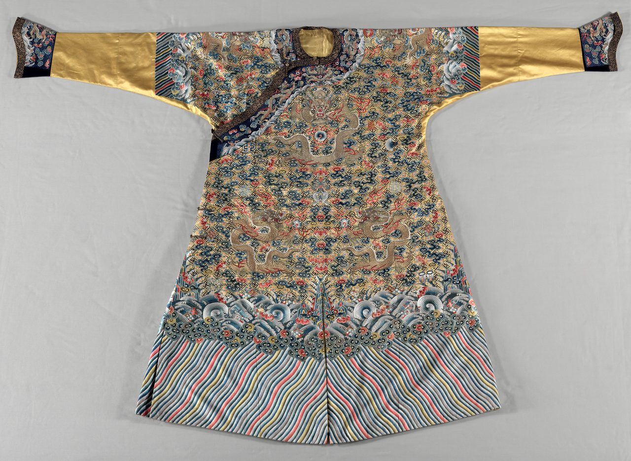 A Qing court robe laid out flat and photographed from directly above on a plain, lightly creased ground, its sleeves extended straight out to left and right and its hem flaring at the bottom below a small round neck opening. The garment is densely embroidered throughout: a field of small scrolling cloud forms with dragons worked among them across the chest and body, a band of stylized breaking waves above the hem, and long diagonal ripple lines below it. The sleeves run as plain smooth panels into dark patterned cuffs.