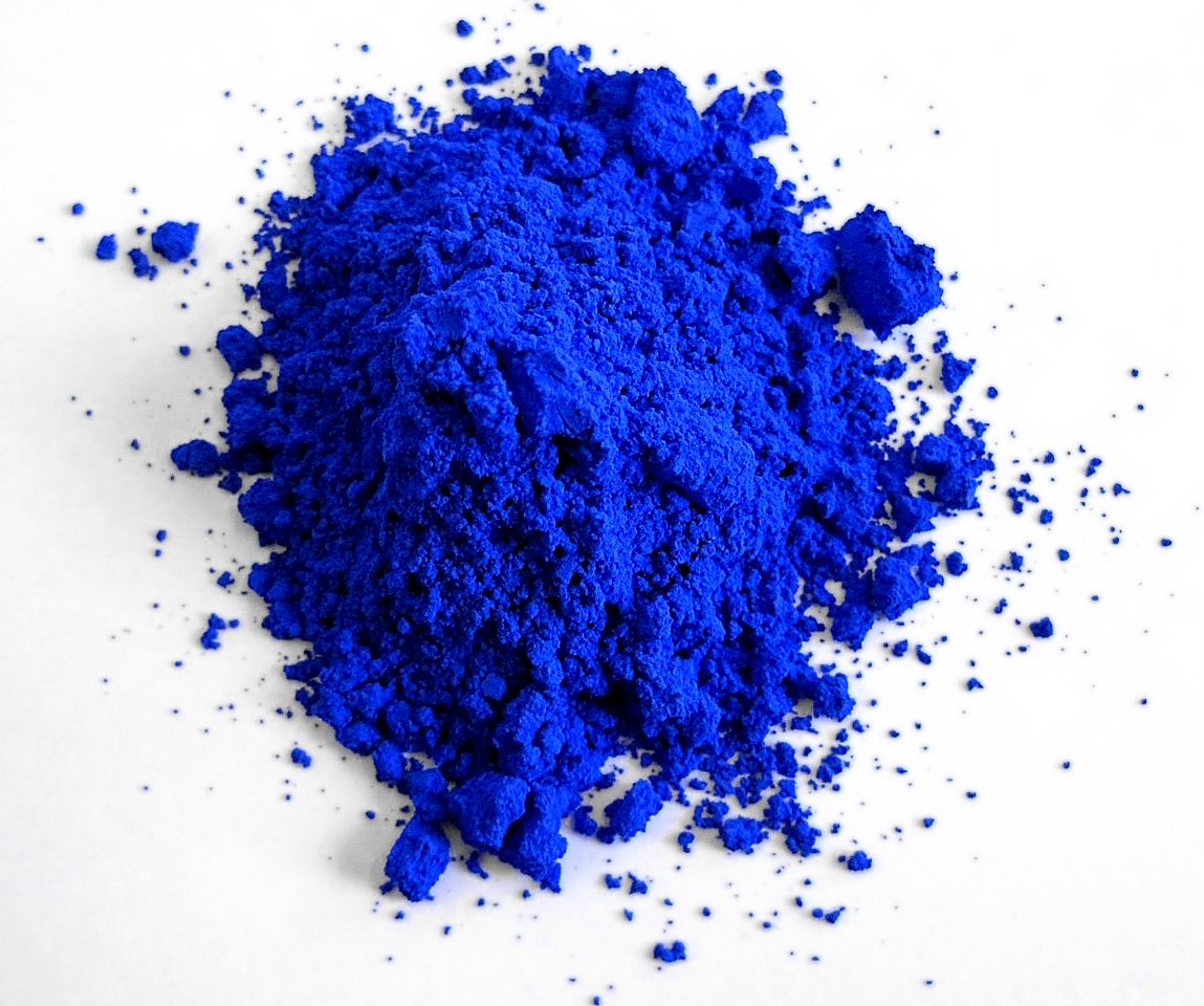 A loose heap of fine powder mounded on a plain white surface and lit from above, its surface broken into small clumps and ridges, with coarser crumbs and single grains scattered outward across the surface all around the pile.