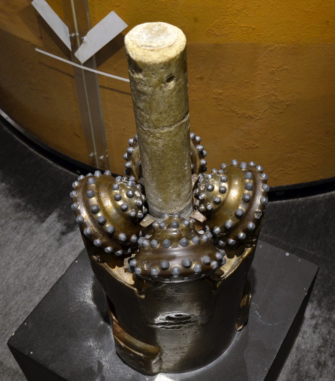 A three cone roller drill bit standing on a black plinth in a museum gallery, its three cutting cones ringed with rows of blunt studs and a pale rough cylinder rising through the centre of them