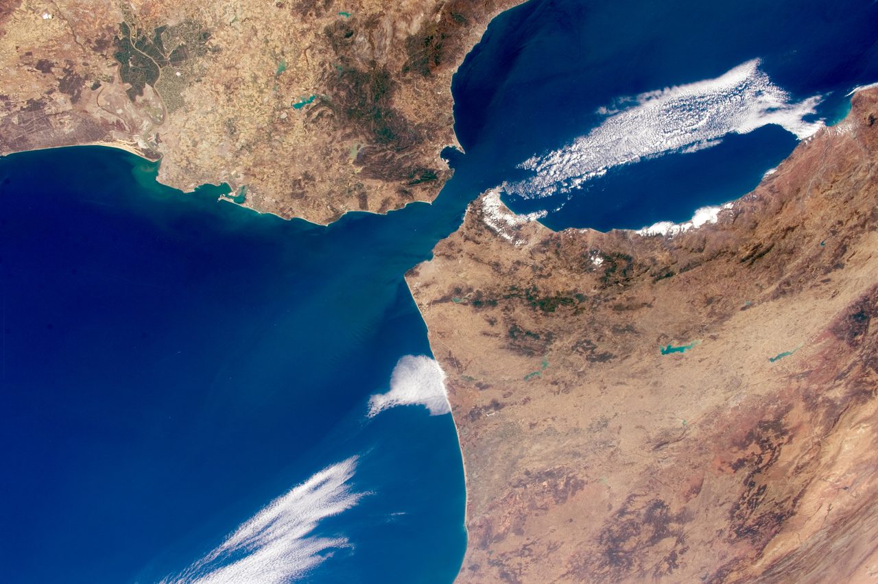 A photograph of the Strait of Gibraltar from orbit, deep blue ocean on one side and a narrower sea on the other, two tan and green coastlines almost meeting at a narrow neck of water, with long streaks of white cloud