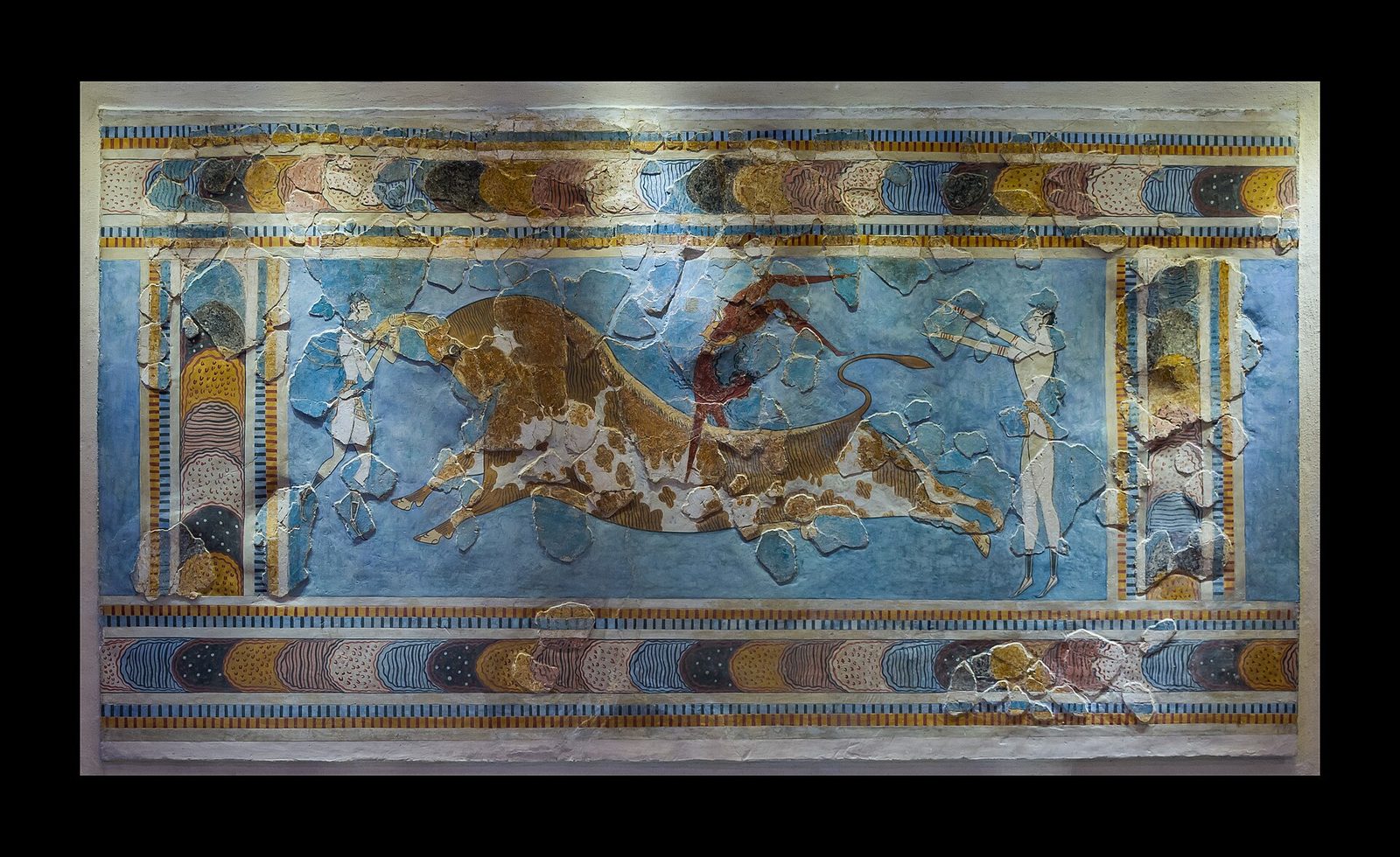 A fresco panel mounted on a wall. A long spotted bull stretches across the centre at full gallop. A red-brown-skinned figure is inverted above its back in mid-vault, a pale-skinned figure grips the horns at the head, and another pale figure stands with arms out behind the tail. A patterned border of coloured discs and bands frames the scene. Many irregular pieces of the painting are raised and hard-edged with crack lines around them, and the surface between those pieces is flat and smooth.