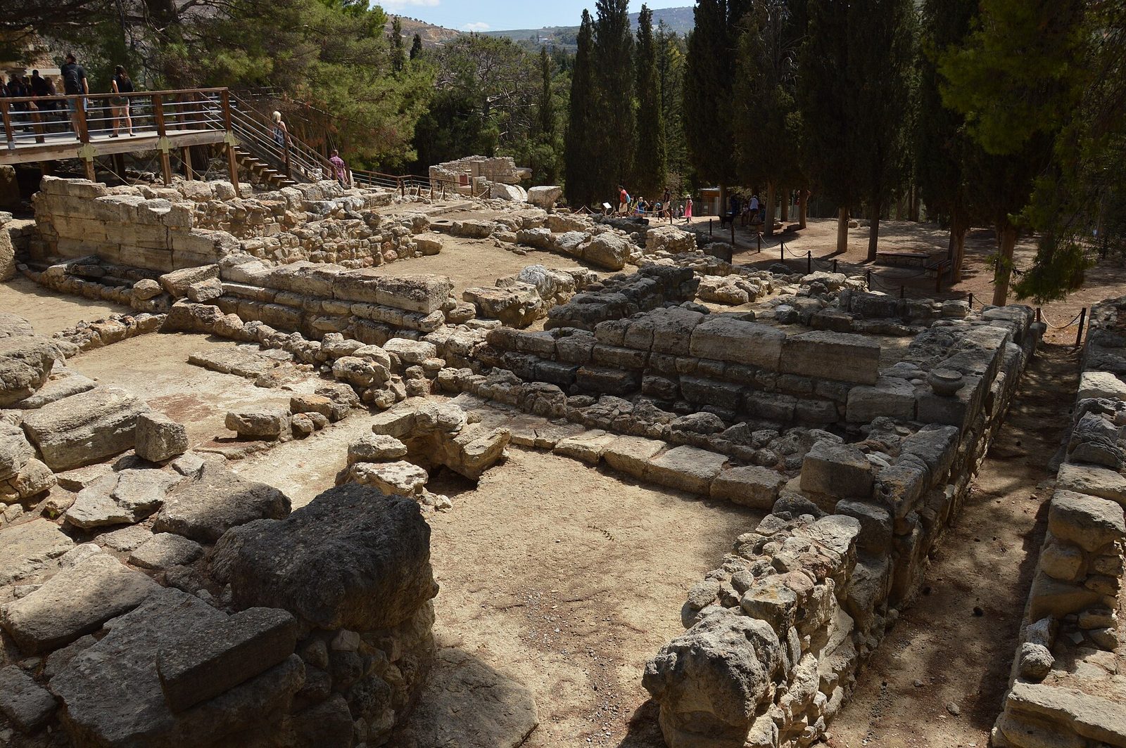 An excavated site of pale stone foundations, low wall stubs and thresholds laid out in rectangular compartments across a slope. Rope barriers on posts run between them, a raised timber viewing walkway crosses the upper left with visitors on it, and dark cypresses and green hillside rise beyond.