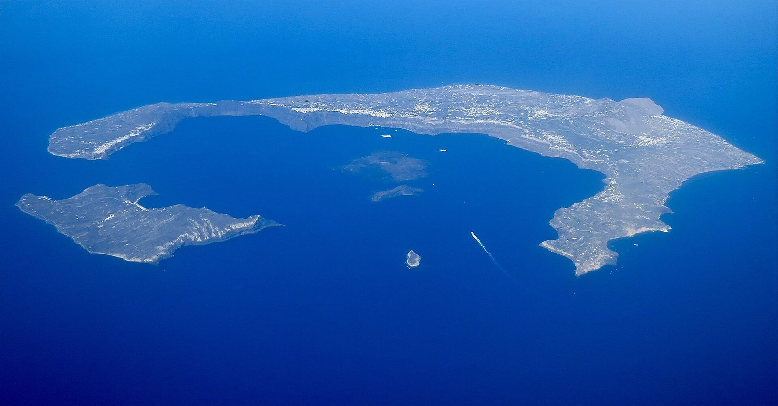 An aerial view of a broken ring of pale, cliff-edged islands enclosing a wide basin of deep blue water. Small dark islets sit near the centre of the basin. A thin white wake trails behind a vessel crossing the water, and open sea fills the distance.