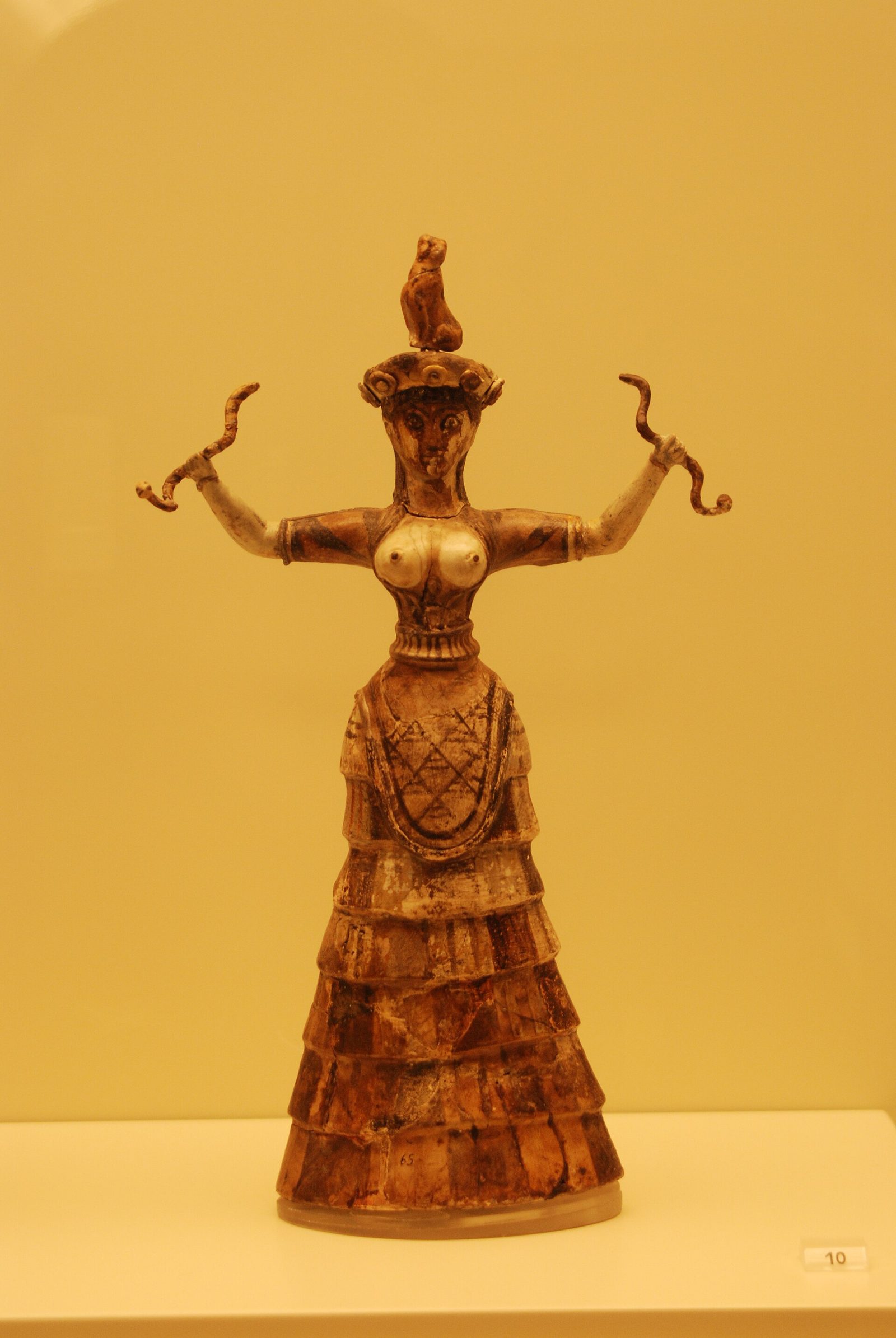 A small standing figurine in a museum case against a warm yellow ground. The female figure holds a snake in each raised, bent arm. She is bare breasted above a tightly laced bodice and wears a long skirt of stacked horizontal flounces. A small seated animal sits on the flat cap on her head.