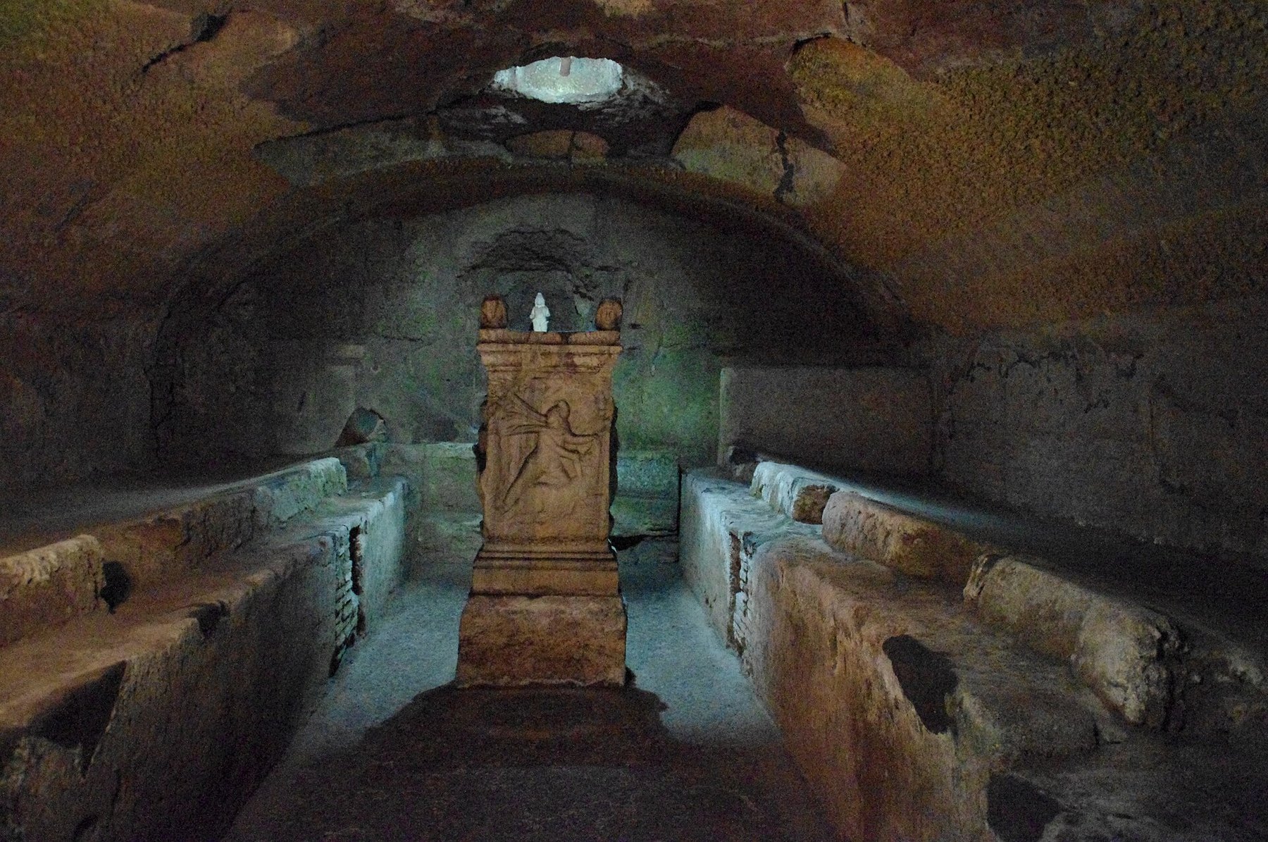 A long, narrow, barrel-vaulted underground chamber in dark stone, dimly lit and mottled brown, ochre and green. Raised stone benches run the full length of both side walls with a floor aisle between them. At the far end stands a freestanding altar block carrying a carved relief of a figure kneeling over an animal. Behind and above it a niche in the wall holds a small pale figure. A light well in the vault at the rear admits a patch of white daylight.