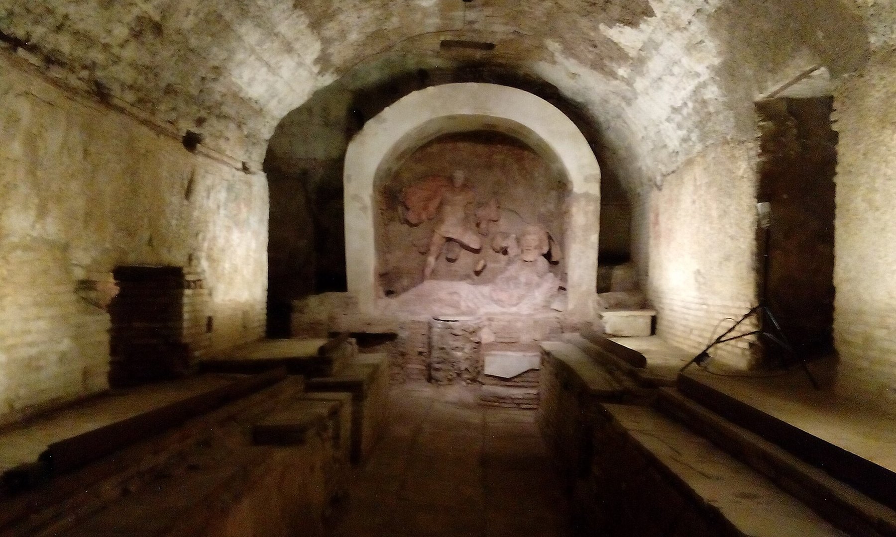 A barrel-vaulted underground chamber in pale stone and brick, warmly lit. At the far end an arched apse holds a stucco relief group of a figure in a billowing cloak over a bull, with traces of red and ochre pigment still on the cloak. A rough masonry altar platform sits below it. Long low benches run along both side walls with a central aisle. Modern lighting equipment, a stand and a cable, stands at the right.
