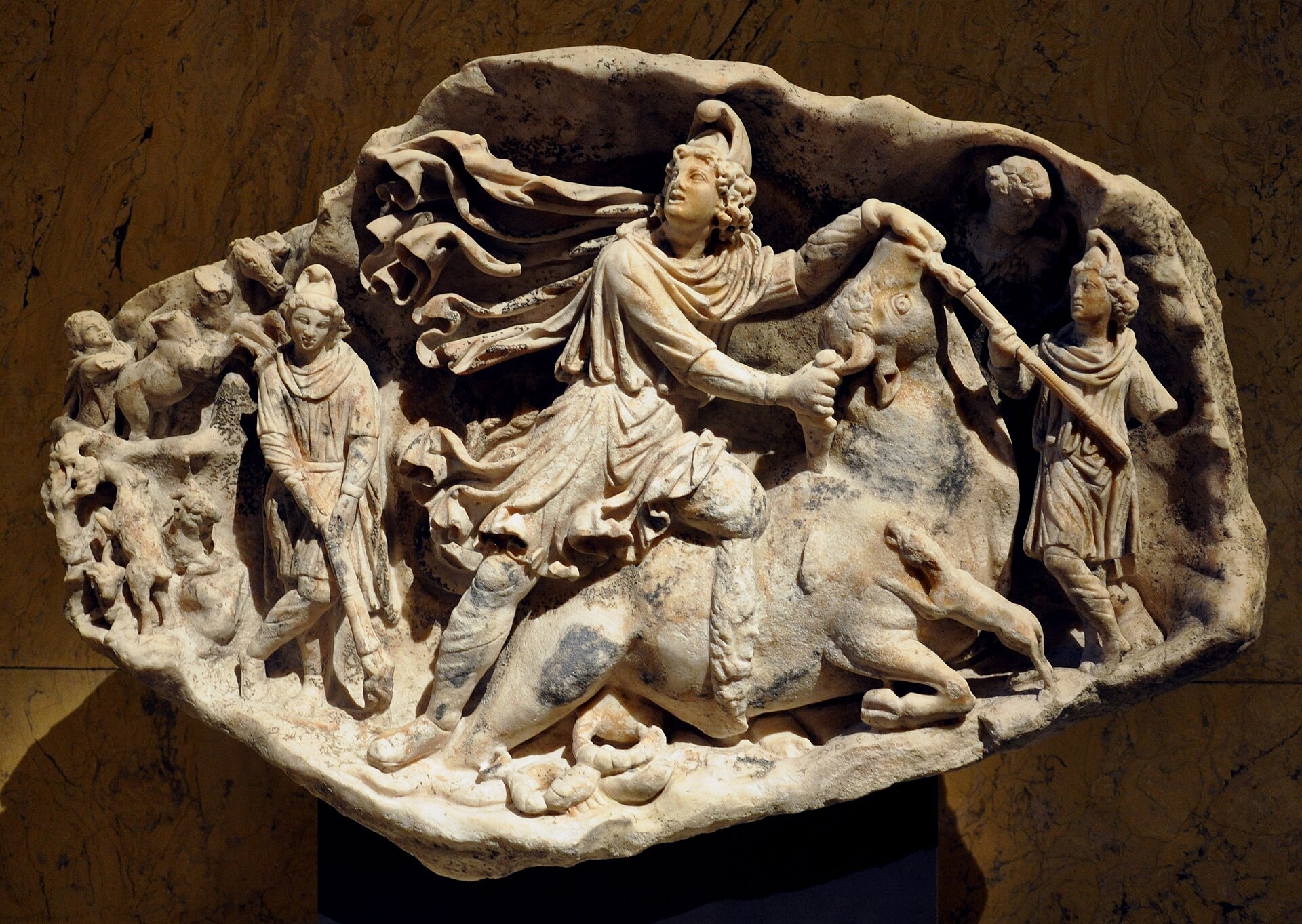 A deep marble relief mounted against a mottled ochre wall. The whole panel is carved as a rough rocky arch, framing the scene like a cave. At the centre a young man in a Phrygian cap with a large billowing cloak kneels on the back of a collapsing bull, his left hand pulling the bull's muzzle up and his right holding a dagger at its neck, his head turned back and away from the animal. A dog leaps at the bull's neck and a snake runs along the ground line beneath. Two smaller figures flank the scene, both in Phrygian caps and short cloaks, each holding a torch, the left one lowered and the right one raised. A vertical column of small subsidiary scenes runs down the left edge.