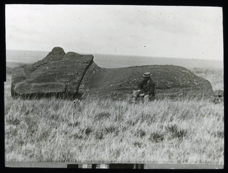 A black-and-white 1914-1915 photograph of a fallen moai lying on what is recorded as one of the ancient transport roads, with a seated man for scale