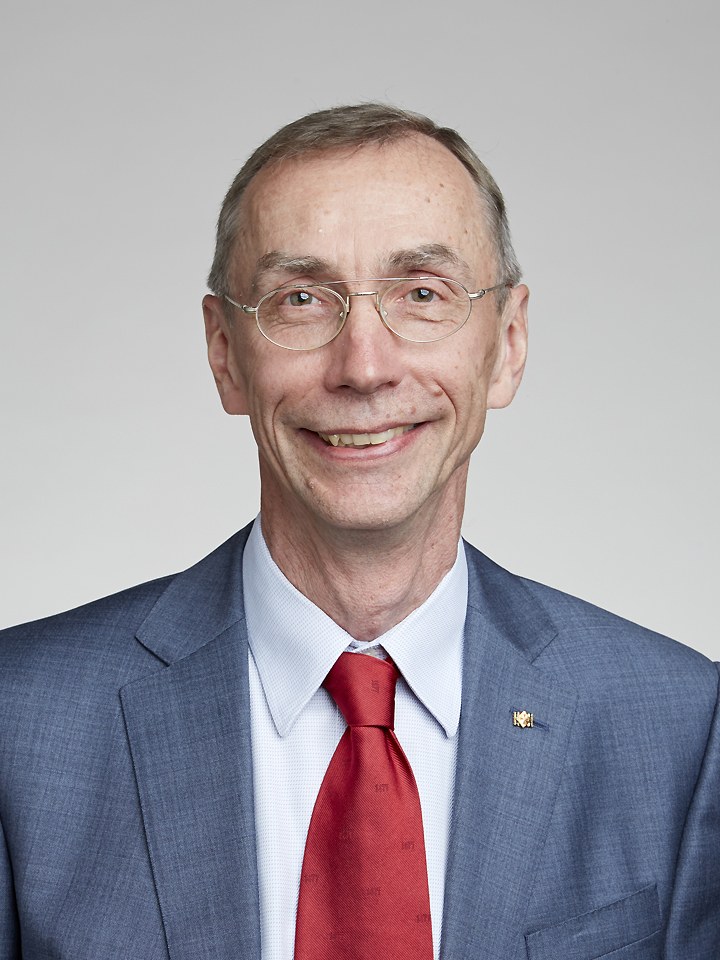 A studio portrait of the geneticist Svante Paabo, an older man with short greying hair and oval wire-rimmed glasses, smiling, wearing a blue-grey suit jacket, pale blue shirt, and a red tie against a neutral grey background