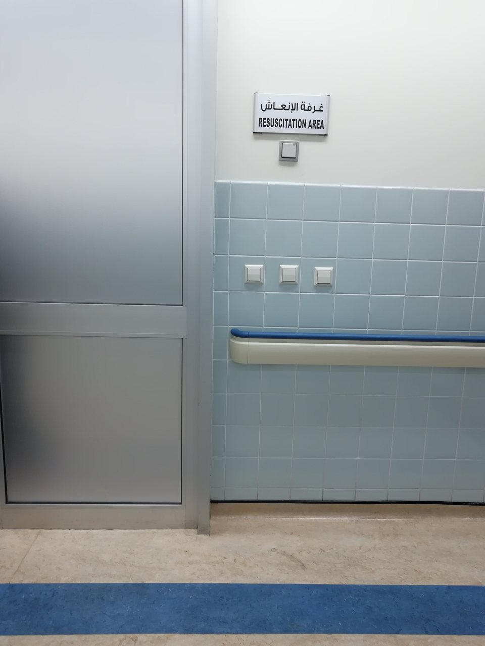 A bilingual hospital wall sign reading RESUSCITATION AREA in Arabic and English, above a blue-tiled wall with a metal door and a corridor floor