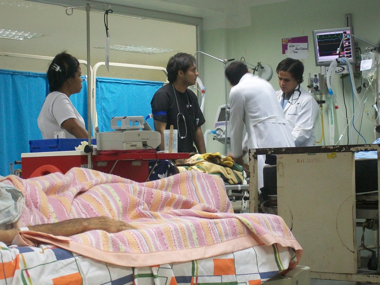 A busy hospital emergency ward with a patient resting under blankets in the foreground and medical staff attending among monitors and equipment