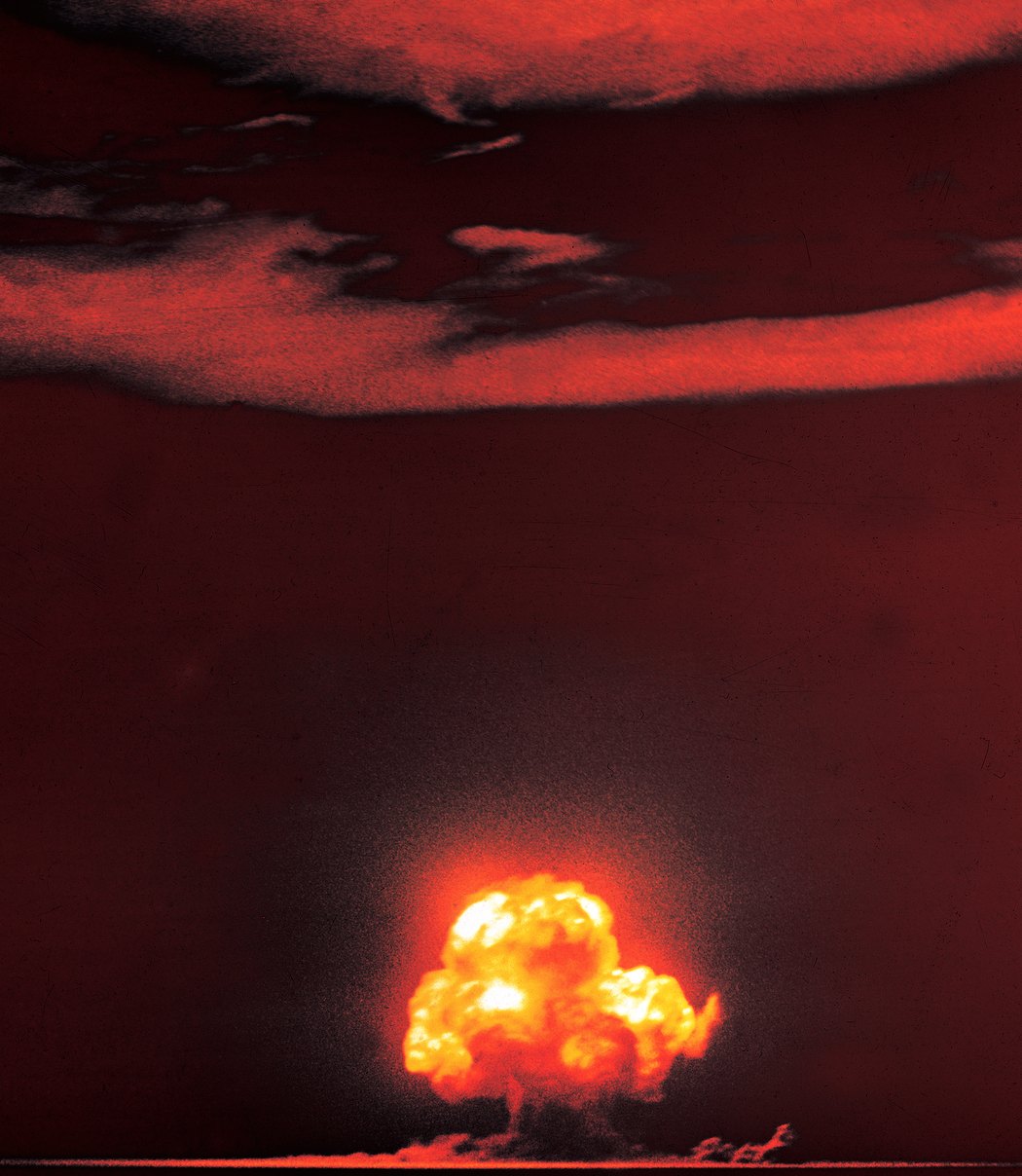 A color photograph of the Trinity test: the roiling orange-and-red fireball of the first nuclear detonation swelling above the New Mexico desert milliseconds after the explosion