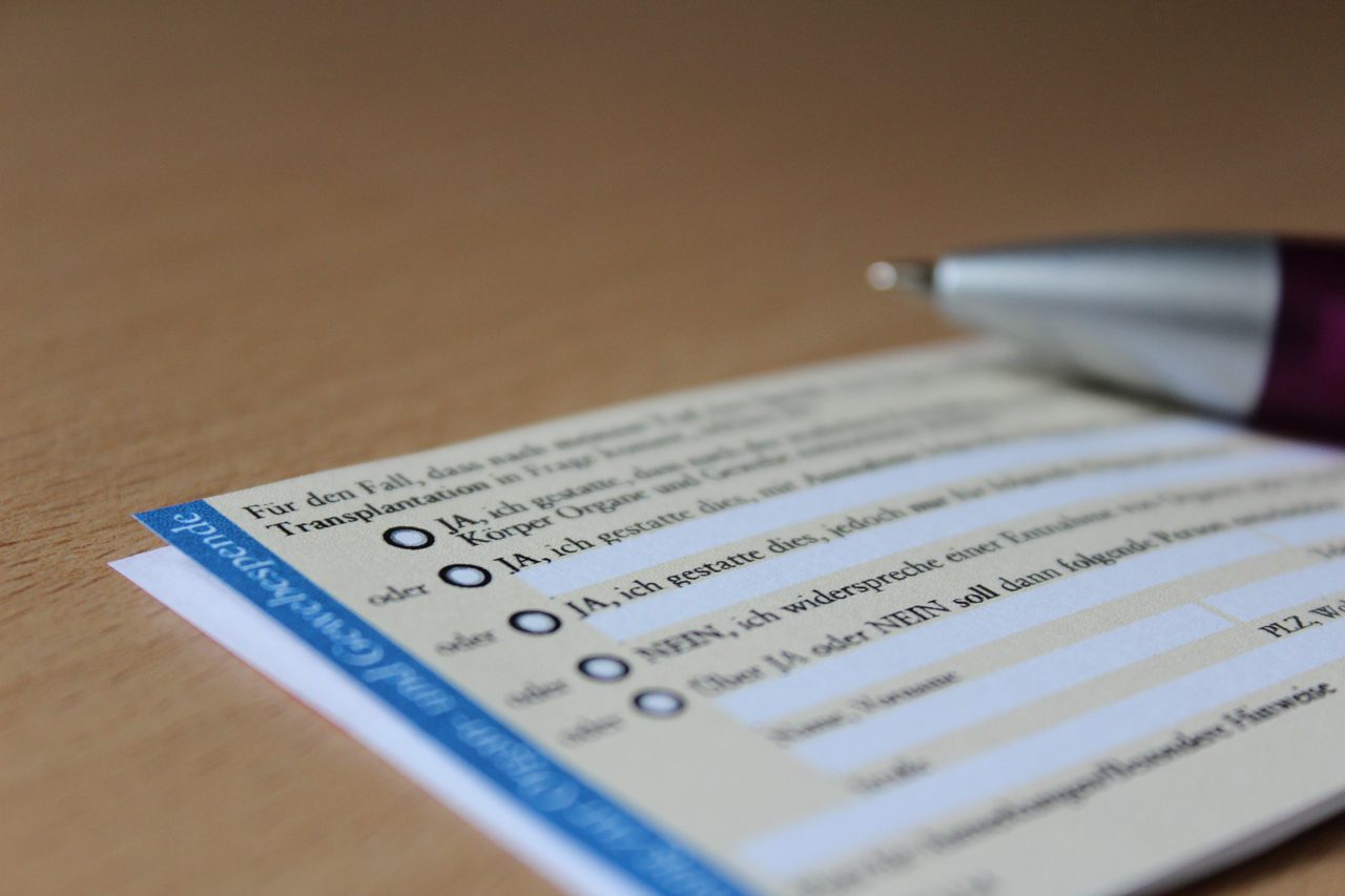 A close up photograph of a printed card lying at a shallow angle on a pale wood surface, running from the lower left of the frame toward the right edge, with a blue band and white lettering along its near side and a column of small circles beside lines of German print across its face, most of it falling out of focus; a ballpoint pen with a silver tip and a dark wine red barrel rests above the card at the upper right, also out of focus.