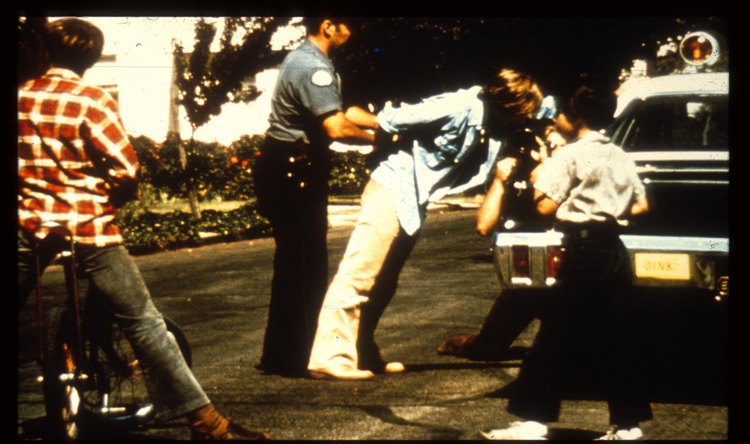 A 1971 color photograph from Philip Zimbardo's Stanford Prison Experiment: a uniformed police officer stages the mock arrest of a young man against a patrol car at the start of the study, watched by bystanders