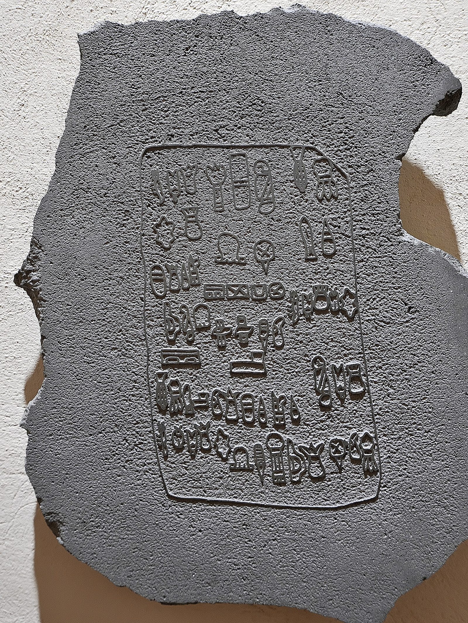 An irregular slab of grey stone with a rough pitted surface, mounted against a white textured wall and casting a shadow. An incised rectangular panel in the centre holds roughly sixty small symbols arranged in rows: hand-like shapes, insect or maize-like forms, ovals, chevrons and banded rectangles.