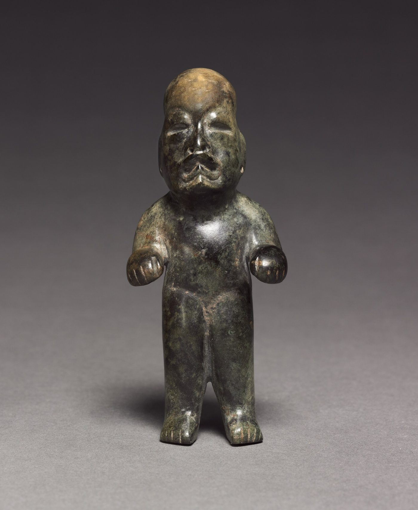 A small standing greenstone figure on a plain graduated grey ground in a sharp studio photograph. It is dark mottled green with a brown patina over the crown, with a broad flattened face, narrow slit eyes, a wide downturned mouth with an everted upper lip, a domed bald head, short arms with fists held forward at the waist, stocky legs and incised toes.