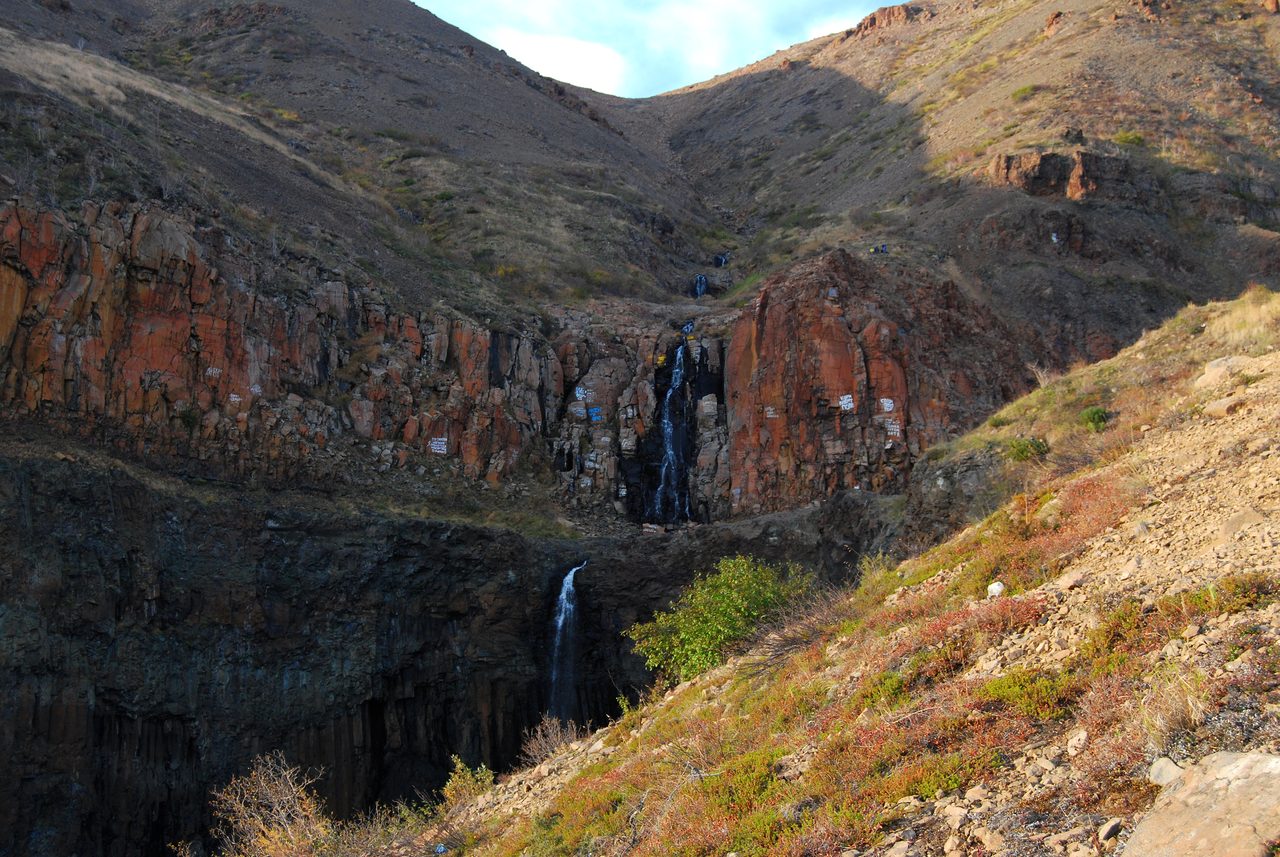 A gorge cut through the Siberian Traps at Krasnye Kamni near Norilsk, with reddish and dark columnar basalt cliffs and a thin waterfall running down the rock face