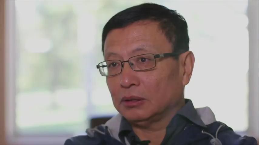 A photograph of the mathematician Yitang Zhang, a middle-aged man with glasses in a grey collared shirt, seated in an interview setting