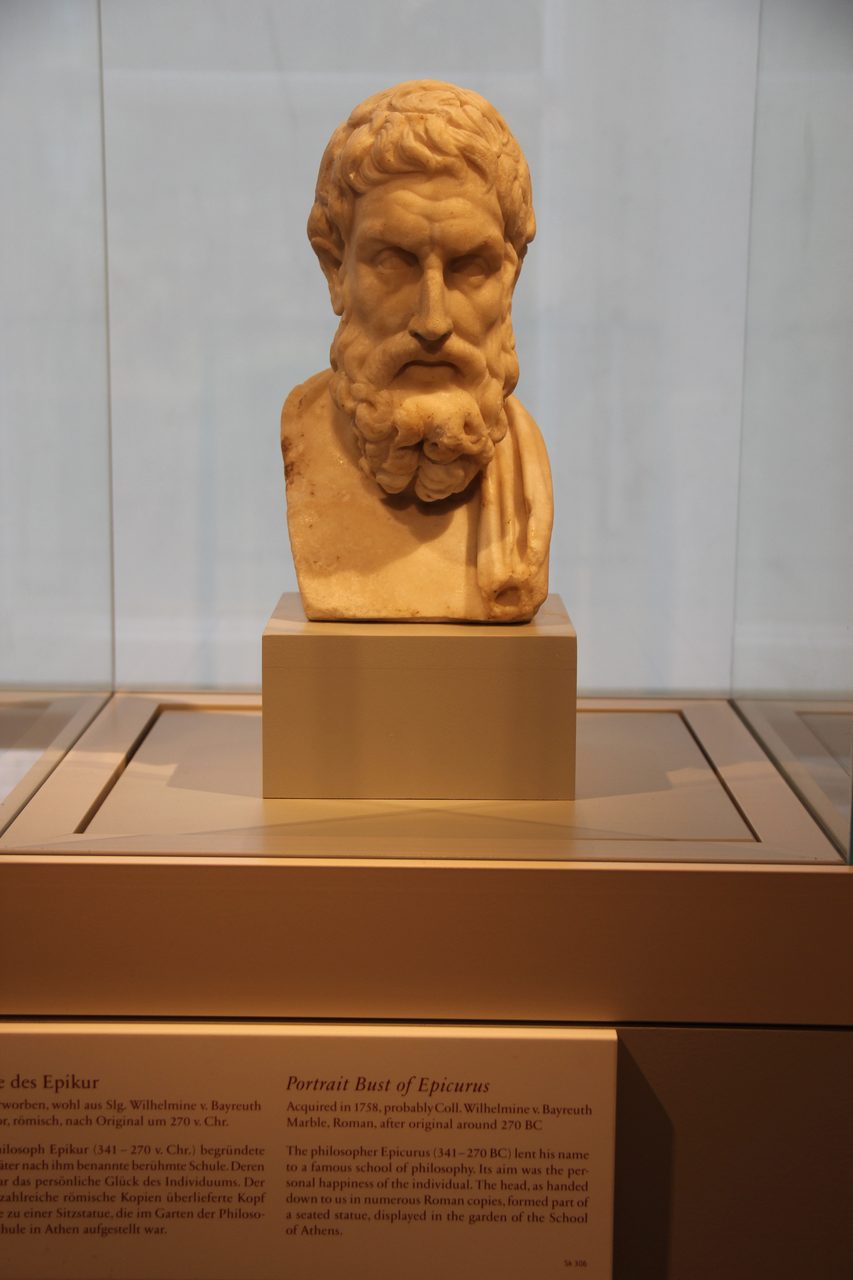 A marble bust of Epicurus on a plinth inside a lit museum vitrine, with the gallery's bilingual wall label legible across the foot of the frame