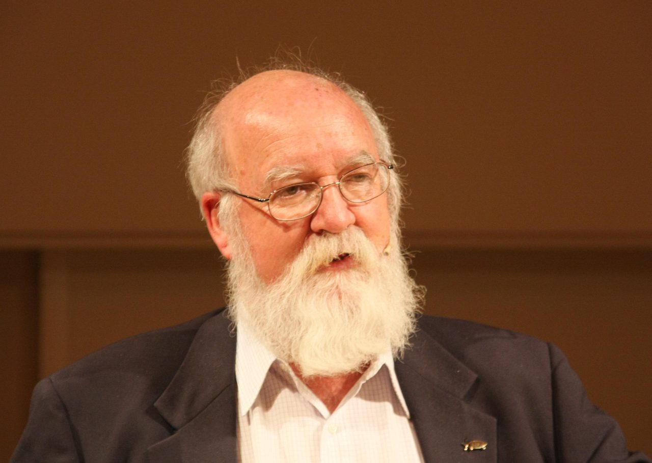 A color photograph of Daniel Dennett speaking, white beard, wire-rimmed glasses, dark jacket over an open-necked check shirt, against a warm brown wall