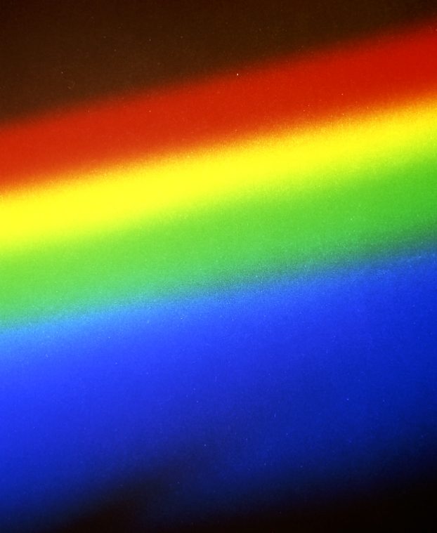 A close photograph of a spectrum cast onto a grainy surface, broad diagonal bands running from deep red through yellow and green into blue against a dark surround