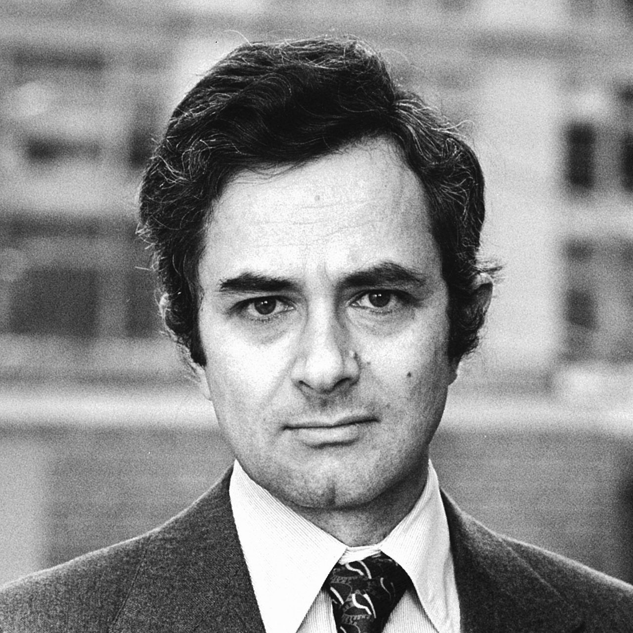A black and white portrait of Thomas Nagel as a younger man, dark hair, jacket and patterned tie, photographed outdoors against a blurred building facade