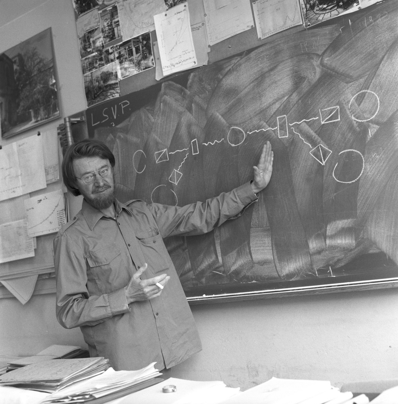 A black-and-white photograph of the physicist John Stewart Bell, bearded and wearing glasses, his dark hair swept back, gesturing in front of a blackboard covered in chalk diagrams