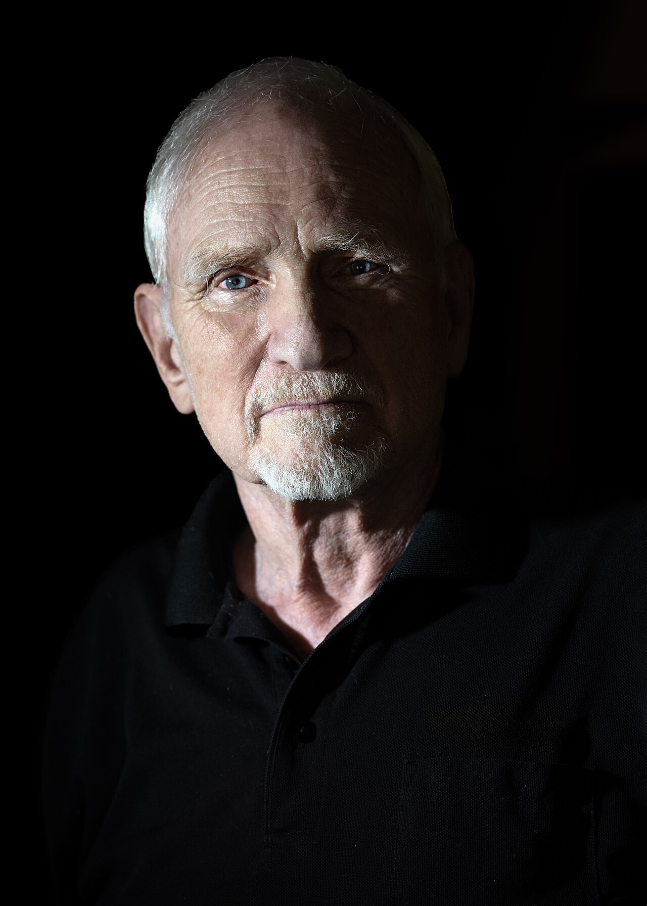 A dramatically lit portrait of the physicist John Clauser as an older man, bald with white hair at the sides and a short white beard, against a black background