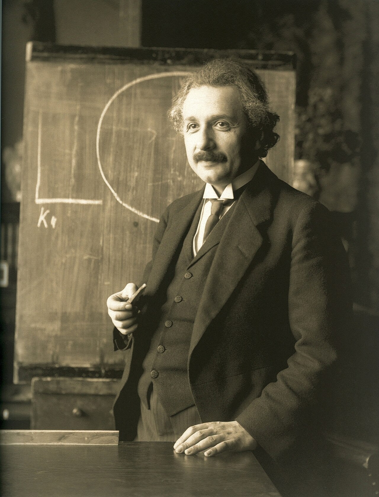 A sepia photograph of a younger Albert Einstein in a three-piece suit, holding a piece of chalk and standing before a chalkboard on which a large loop and the letter K are drawn