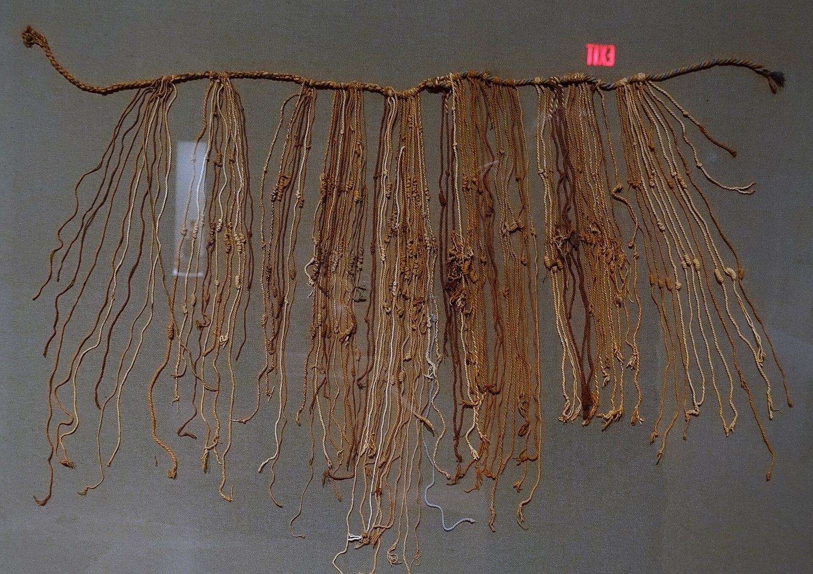 A real Inca quipu displayed against a grey-green backing: a horizontal main cord with dozens of tan and brown pendant cords hanging down from it, each bearing knots at various positions along its length