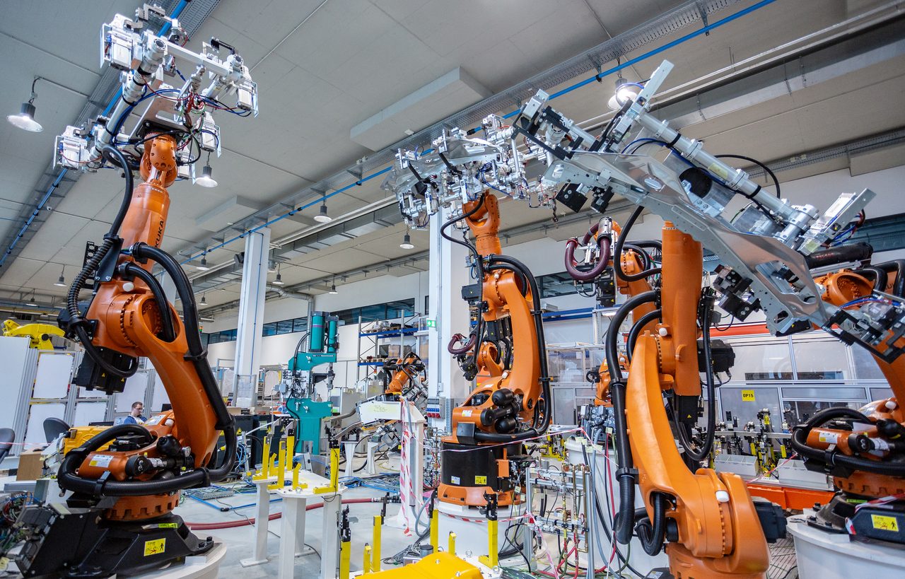 A row of industrial robot arms working along an automated production line inside a factory hall, behind safety guarding