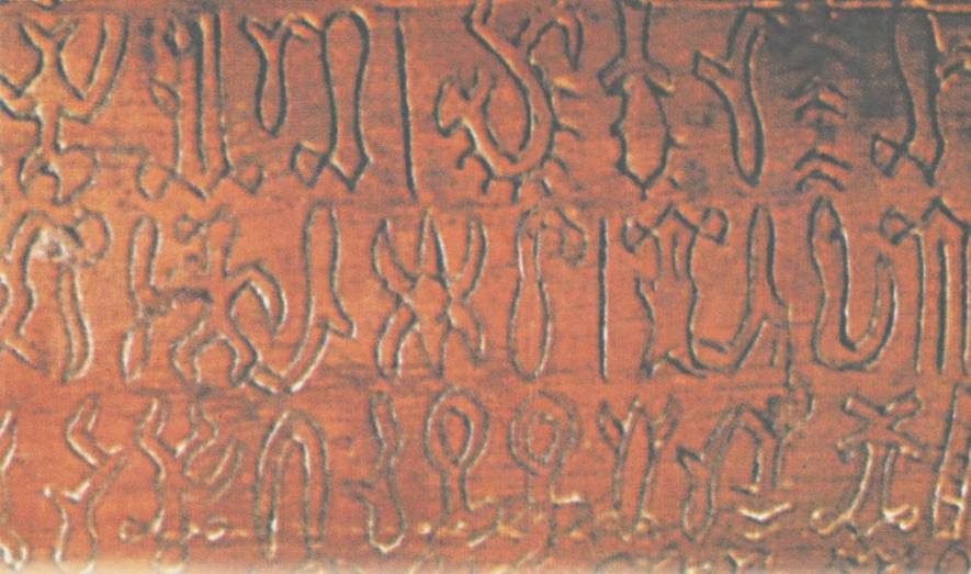 A close colour view of a reddish-brown wooden surface filling the frame, cut with three horizontal rows of small incised signs. The signs are shallow grooves catching pale light against the darker wood, and read as stylised figures, hooks, crescents and stick-like forms. The wood grain runs horizontally behind them.